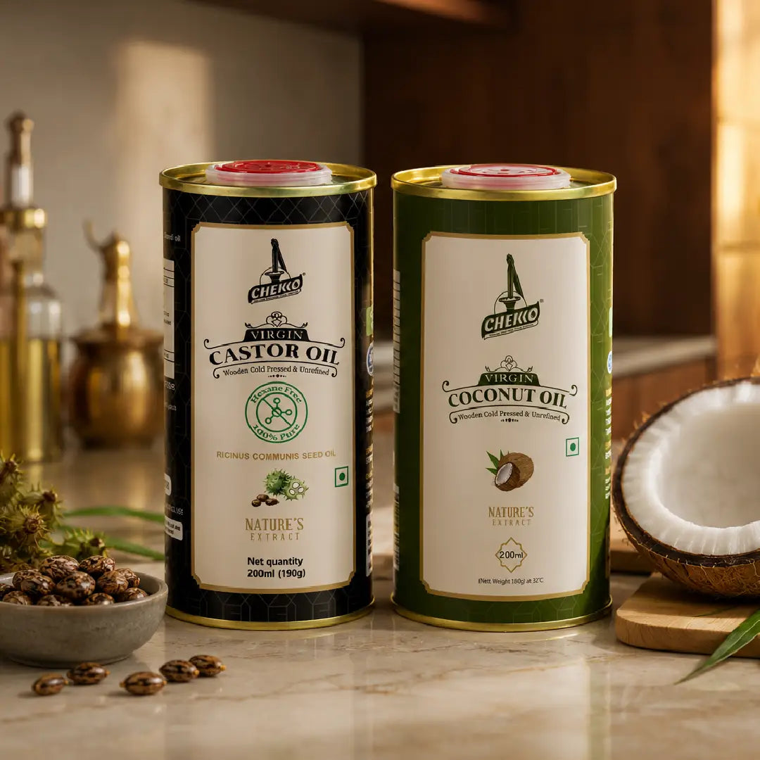 Two tall cylindrical metal tins of Chekko Naturals coconut and castor oil displayed on a countertop with kitchen props, including a halved coconut, seeds, and brass bottles in the background.