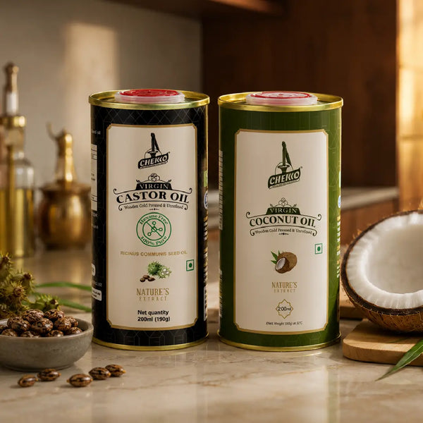 Two tall cylindrical metal tins of Chekko Naturals coconut and castor oil displayed on a countertop with kitchen props, including a halved coconut, seeds, and brass bottles in the background.