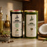 Two tall cylindrical metal tins of Chekko Naturals coconut and castor oil displayed on a countertop with kitchen props, including a halved coconut, seeds, and brass bottles in the background.